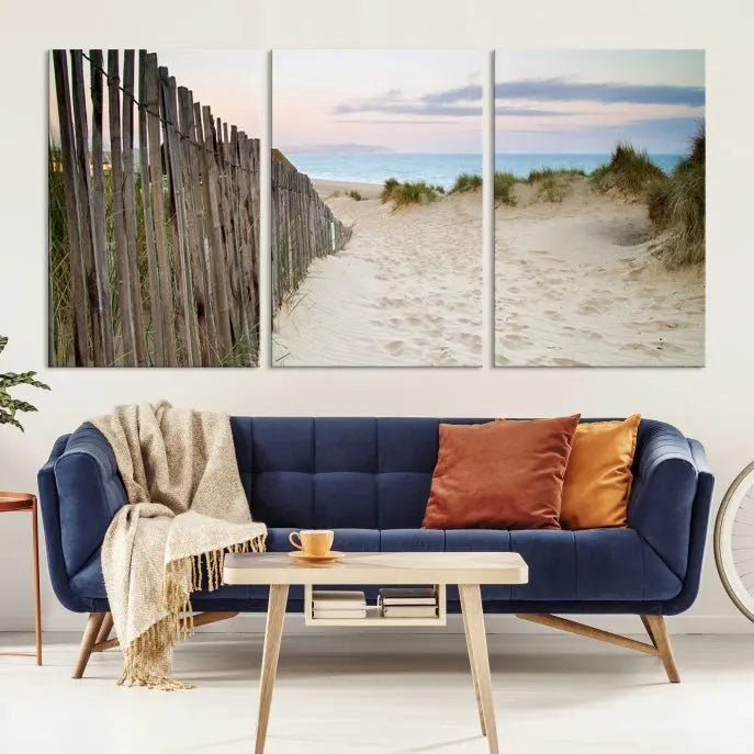 The Coastal Beach Fence Wall Art Ocean Landscape Canvas Print, featuring a triptych of a sandy beach scene with dunes and fences, is gallery wrapped on museum-quality canvas and displayed on a modern dark wall.