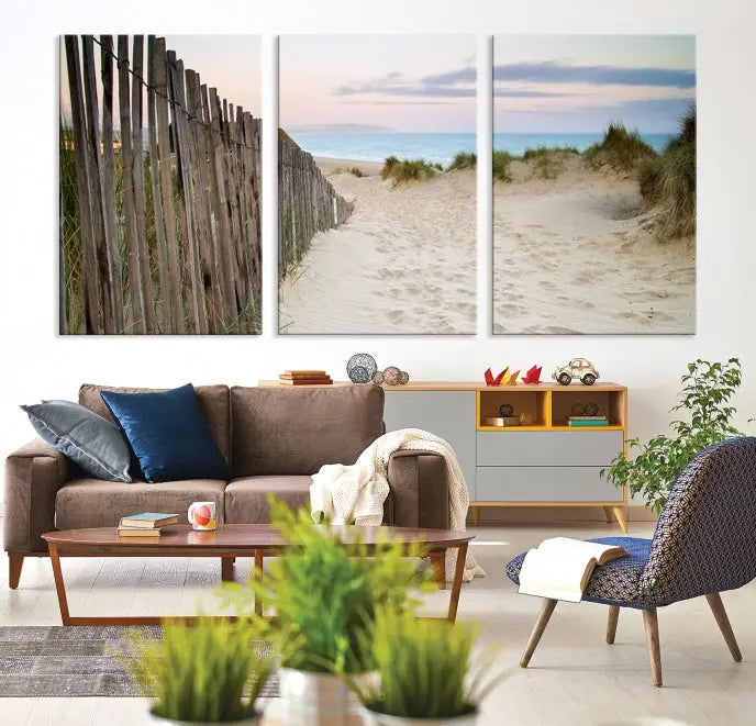 The Coastal Beach Fence Wall Art Ocean Landscape Canvas Print, featuring a triptych of a sandy beach scene with dunes and fences, is gallery wrapped on museum-quality canvas and displayed on a modern dark wall.