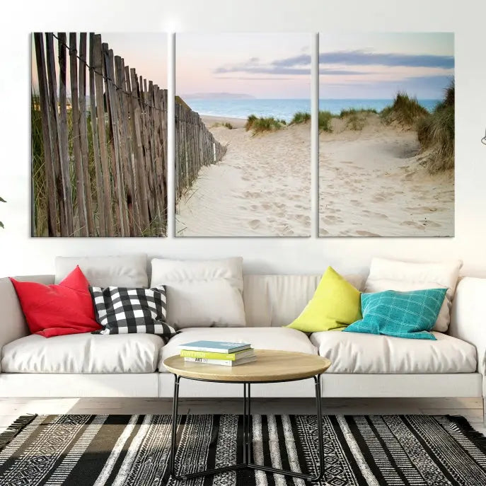 The Coastal Beach Fence Wall Art Ocean Landscape Canvas Print, featuring a triptych of a sandy beach scene with dunes and fences, is gallery wrapped on museum-quality canvas and displayed on a modern dark wall.