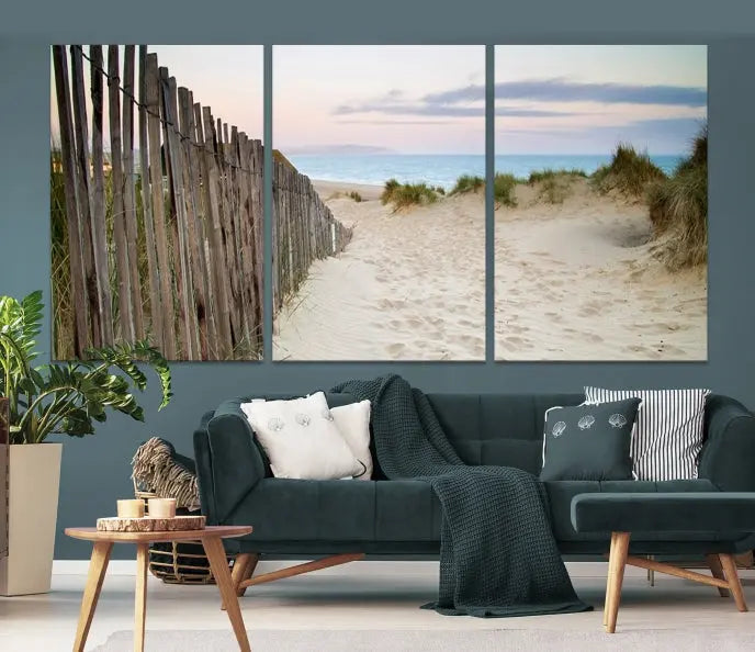 The Coastal Beach Fence Wall Art Ocean Landscape Canvas Print, featuring a triptych of a sandy beach scene with dunes and fences, is gallery wrapped on museum-quality canvas and displayed on a modern dark wall.