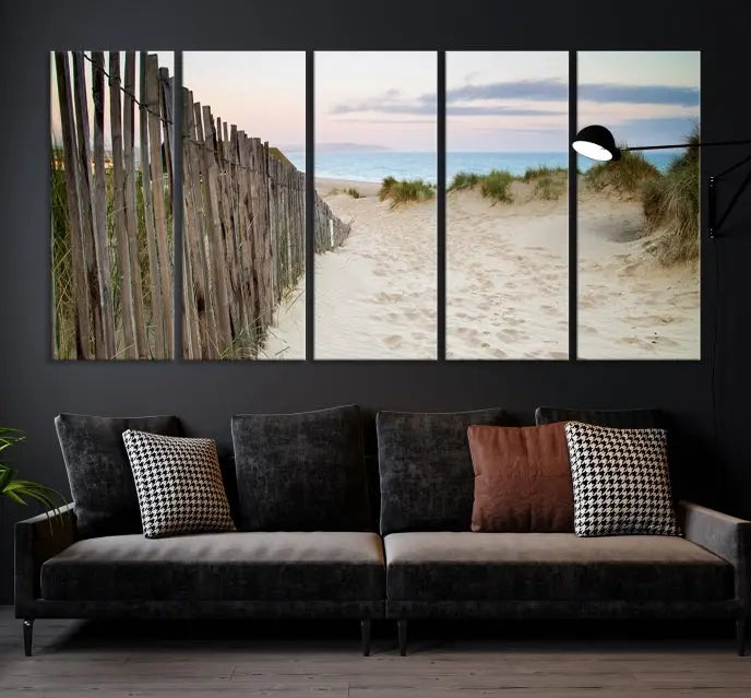 The Coastal Beach Fence Wall Art Ocean Landscape Canvas Print, featuring a triptych of a sandy beach scene with dunes and fences, is gallery wrapped on museum-quality canvas and displayed on a modern dark wall.