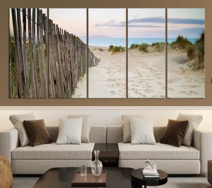 The Coastal Beach Fence Wall Art Ocean Landscape Canvas Print, featuring a triptych of a sandy beach scene with dunes and fences, is gallery wrapped on museum-quality canvas and displayed on a modern dark wall.