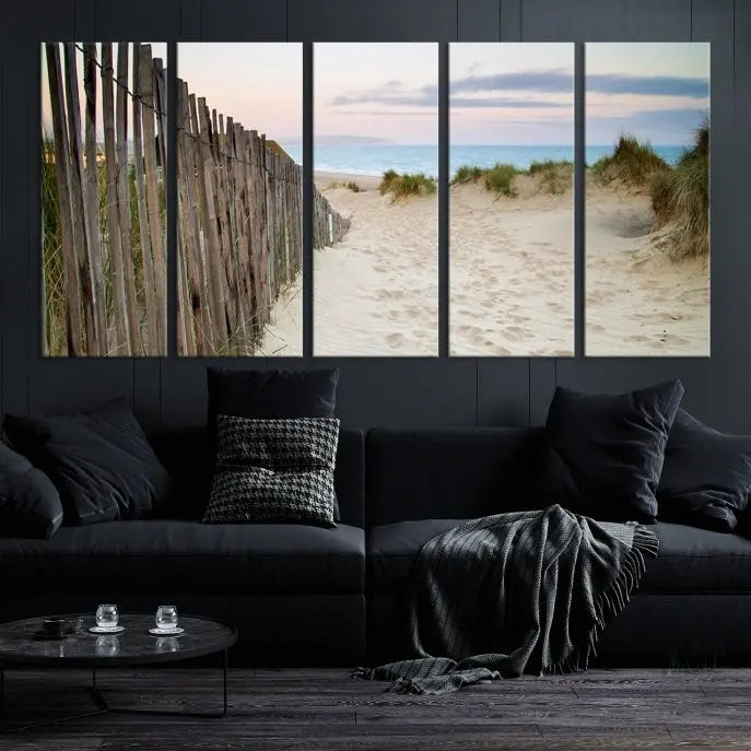 The Coastal Beach Fence Wall Art Ocean Landscape Canvas Print, featuring a triptych of a sandy beach scene with dunes and fences, is gallery wrapped on museum-quality canvas and displayed on a modern dark wall.