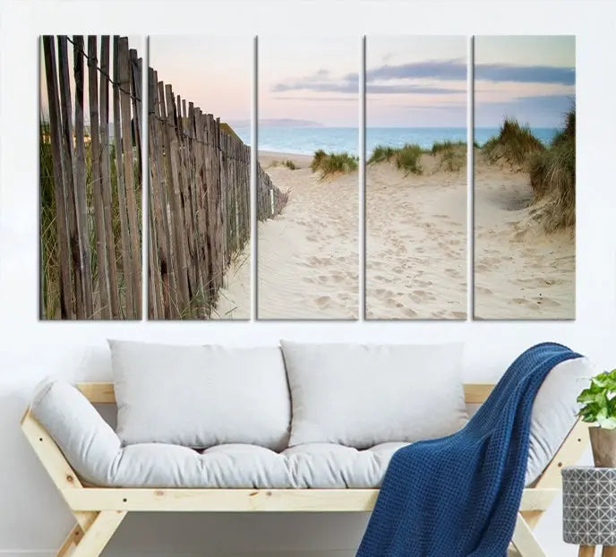 The Coastal Beach Fence Wall Art Ocean Landscape Canvas Print, featuring a triptych of a sandy beach scene with dunes and fences, is gallery wrapped on museum-quality canvas and displayed on a modern dark wall.