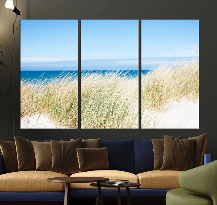 The Coastal Ocean Beach Wall Art Canvas Print, featuring a three-panel design with beach grass and an ocean view on museum-quality canvas, enhances the space in a modern living room. The artwork is ready to hang and comes with a UV-protective coating for enduring beauty.