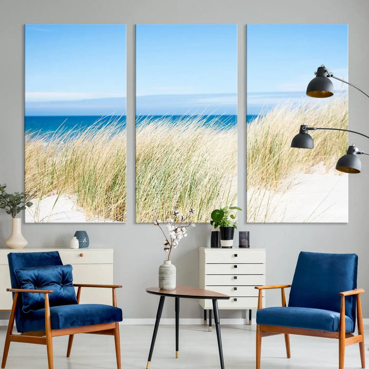 The Coastal Ocean Beach Wall Art Canvas Print, featuring a three-panel design with beach grass and an ocean view on museum-quality canvas, enhances the space in a modern living room. The artwork is ready to hang and comes with a UV-protective coating for enduring beauty.