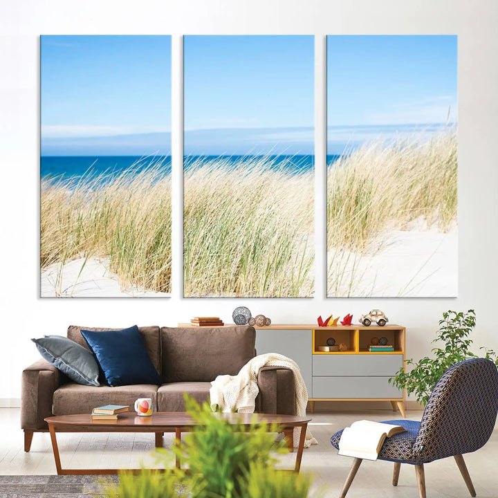 The Coastal Ocean Beach Wall Art Canvas Print, featuring a three-panel design with beach grass and an ocean view on museum-quality canvas, enhances the space in a modern living room. The artwork is ready to hang and comes with a UV-protective coating for enduring beauty.