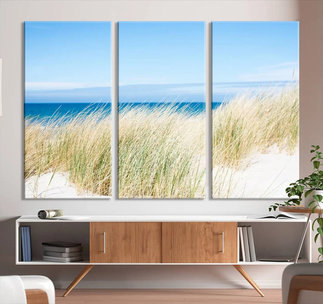 The Coastal Ocean Beach Wall Art Canvas Print, featuring a three-panel design with beach grass and an ocean view on museum-quality canvas, enhances the space in a modern living room. The artwork is ready to hang and comes with a UV-protective coating for enduring beauty.