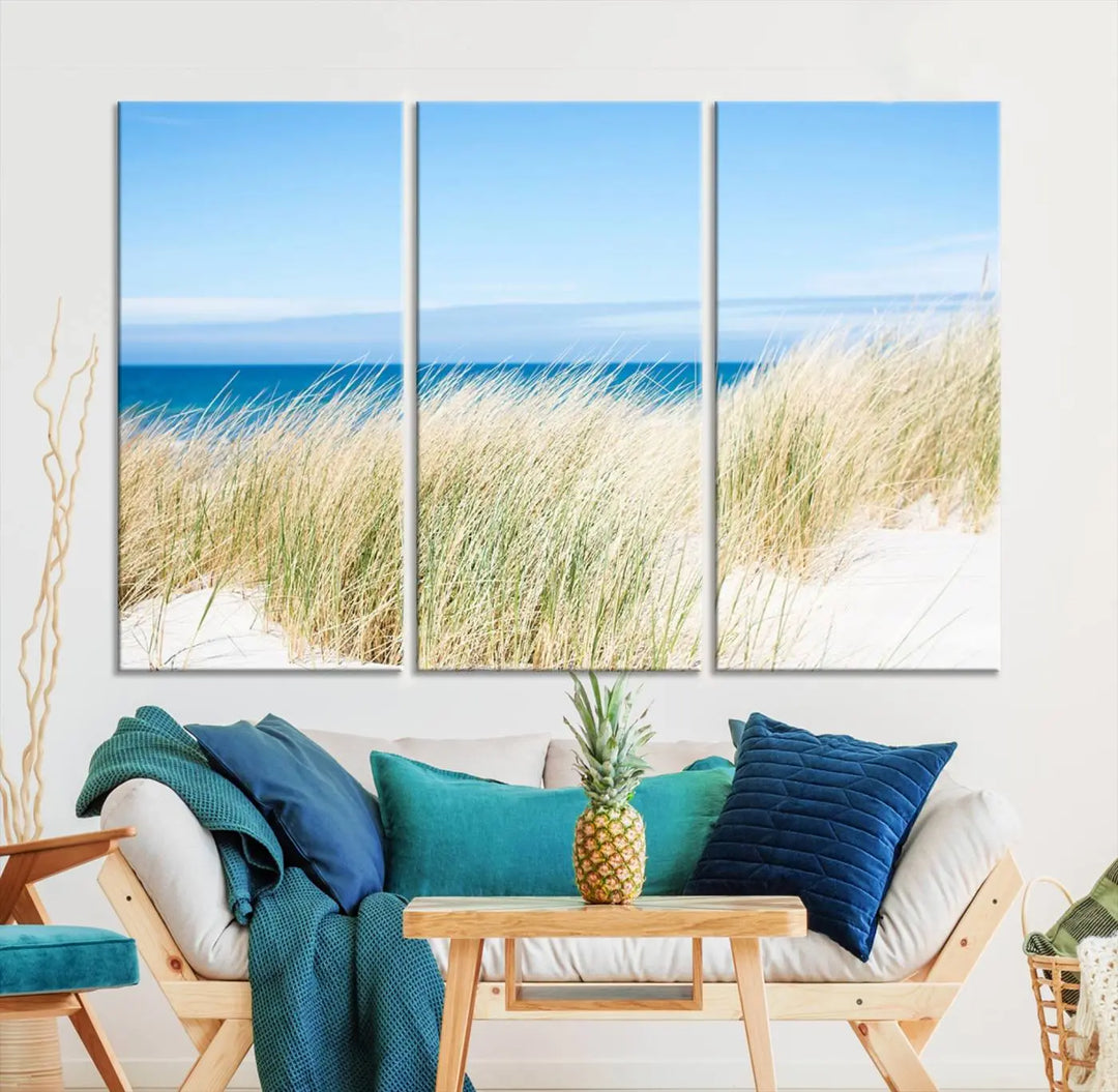 The Coastal Ocean Beach Wall Art Canvas Print, featuring a three-panel design with beach grass and an ocean view on museum-quality canvas, enhances the space in a modern living room. The artwork is ready to hang and comes with a UV-protective coating for enduring beauty.