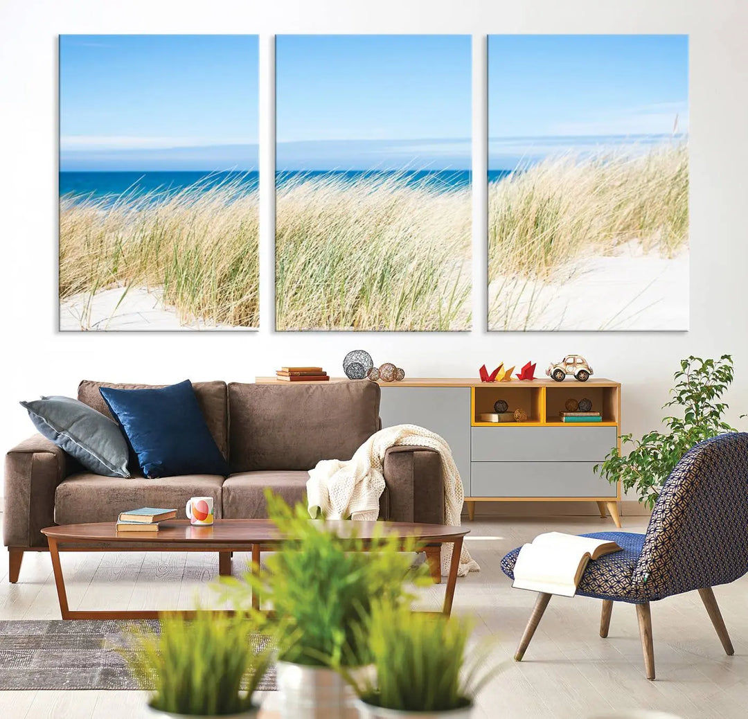 The Coastal Ocean Beach Wall Art Canvas Print, featuring a three-panel design with beach grass and an ocean view on museum-quality canvas, enhances the space in a modern living room. The artwork is ready to hang and comes with a UV-protective coating for enduring beauty.