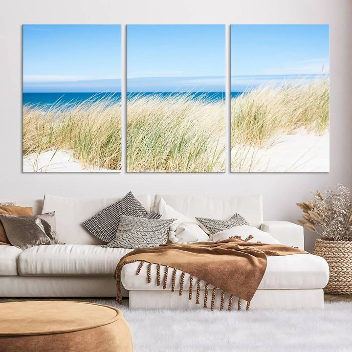 The Coastal Ocean Beach Wall Art Canvas Print, featuring a three-panel design with beach grass and an ocean view on museum-quality canvas, enhances the space in a modern living room. The artwork is ready to hang and comes with a UV-protective coating for enduring beauty.