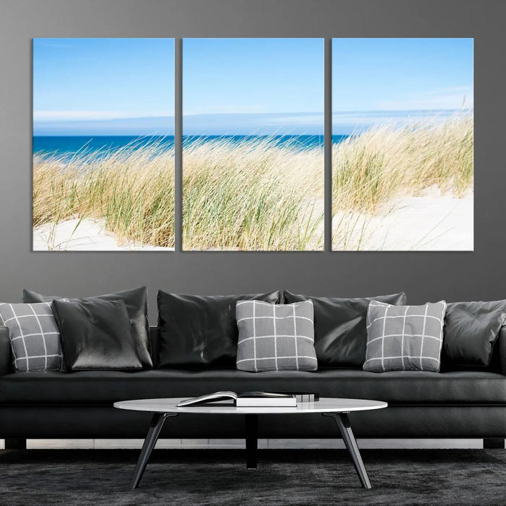 The Coastal Ocean Beach Wall Art Canvas Print, featuring a three-panel design with beach grass and an ocean view on museum-quality canvas, enhances the space in a modern living room. The artwork is ready to hang and comes with a UV-protective coating for enduring beauty.