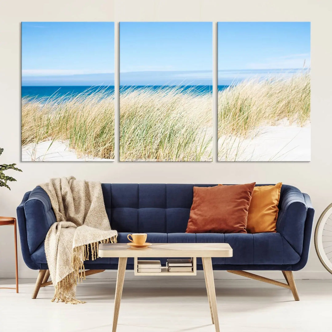 The Coastal Ocean Beach Wall Art Canvas Print, featuring a three-panel design with beach grass and an ocean view on museum-quality canvas, enhances the space in a modern living room. The artwork is ready to hang and comes with a UV-protective coating for enduring beauty.