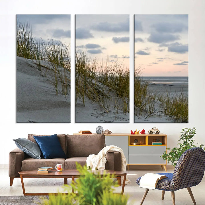 The Coastal Ocean Landscape Beach Wall Art Canvas Print is a triptych depicting sandy dunes and grass at sunset. It arrives ready to hang and is equipped with a UV-protective coating for lasting vibrancy and protection against fading sunlight.