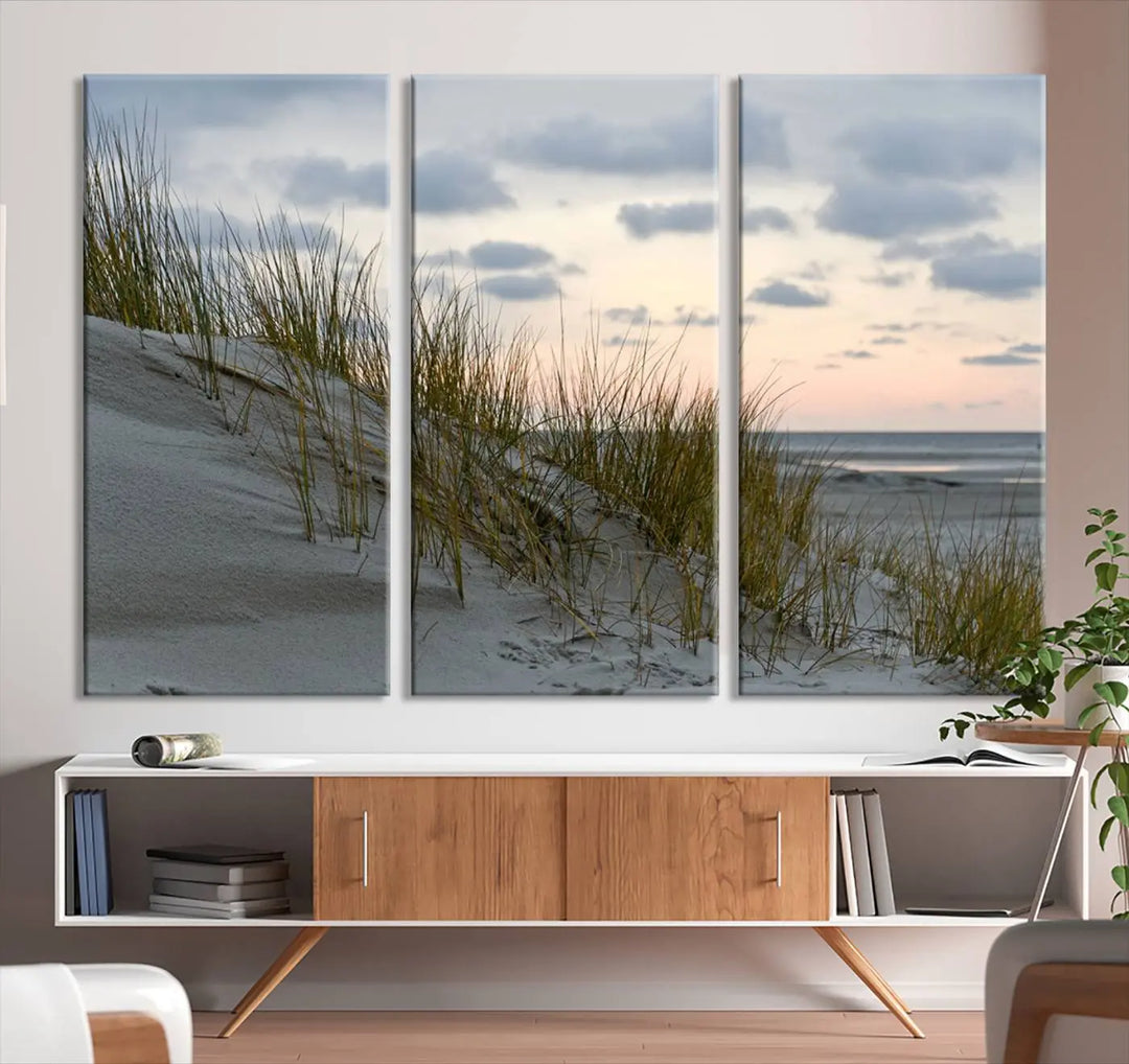 The Coastal Ocean Landscape Beach Wall Art Canvas Print is a triptych depicting sandy dunes and grass at sunset. It arrives ready to hang and is equipped with a UV-protective coating for lasting vibrancy and protection against fading sunlight.