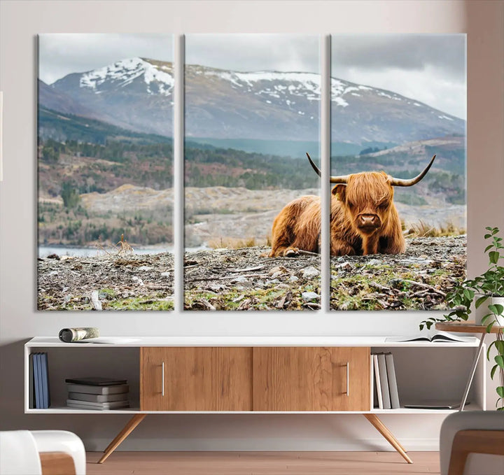 The Highland Cow Horn Farm Wall Art Canvas Print showcases a serene scene of a highland cow resting with snow-capped mountains in the background. This three-panel, museum-quality canvas features a UV-protective coating and is ready to hang.