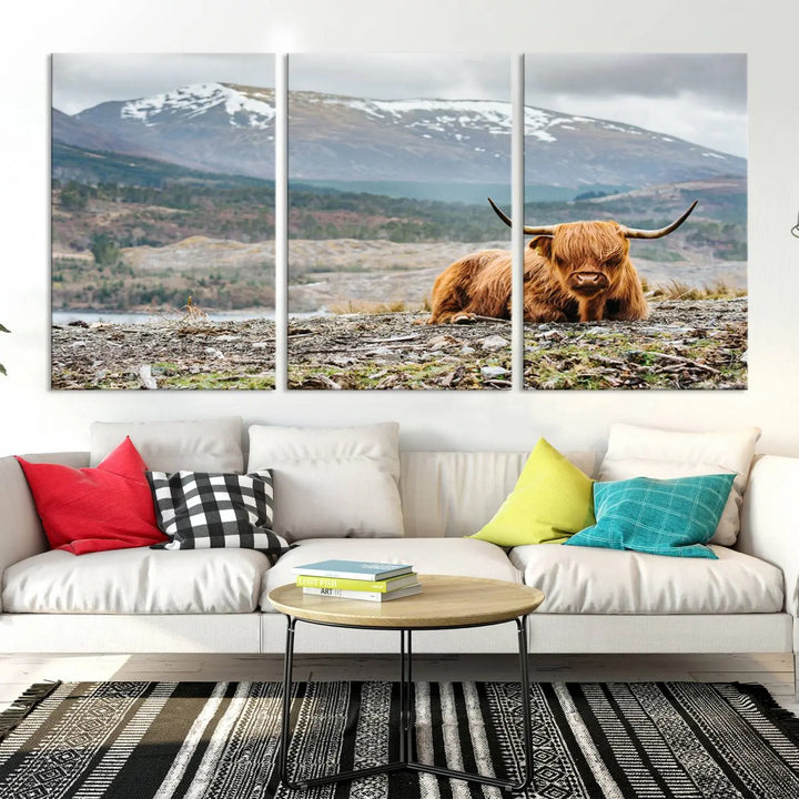 The Highland Cow Horn Farm Wall Art Canvas Print showcases a serene scene of a highland cow resting with snow-capped mountains in the background. This three-panel, museum-quality canvas features a UV-protective coating and is ready to hang.