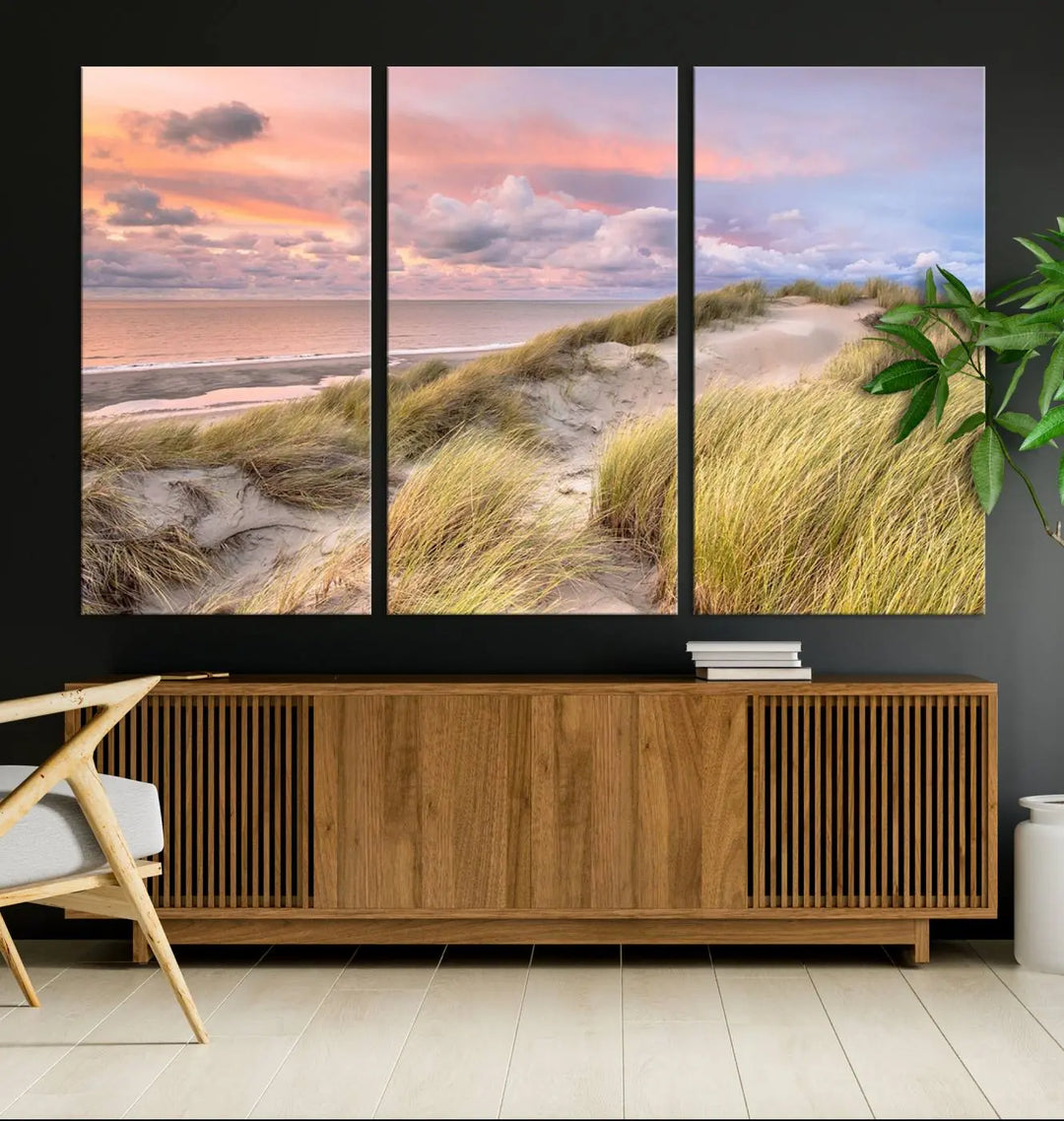 The Ocean Beach Wall Art Canvas Print Sunset Artwork captures a stunning sunset over sand dunes and grass, adding a touch of coastal charm to the room. This piece of wall art infuses any space with warmth and serenity.