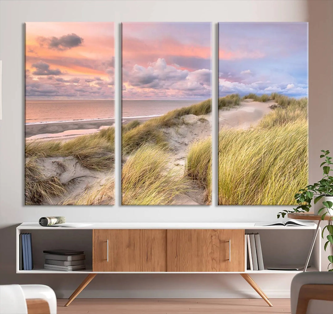 The Ocean Beach Wall Art Canvas Print Sunset Artwork captures a stunning sunset over sand dunes and grass, adding a touch of coastal charm to the room. This piece of wall art infuses any space with warmth and serenity.