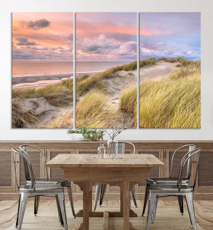 The Ocean Beach Wall Art Canvas Print Sunset Artwork captures a stunning sunset over sand dunes and grass, adding a touch of coastal charm to the room. This piece of wall art infuses any space with warmth and serenity.