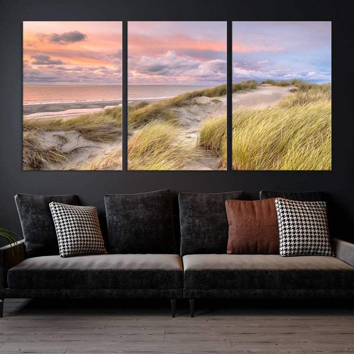 The Ocean Beach Wall Art Canvas Print Sunset Artwork captures a stunning sunset over sand dunes and grass, adding a touch of coastal charm to the room. This piece of wall art infuses any space with warmth and serenity.