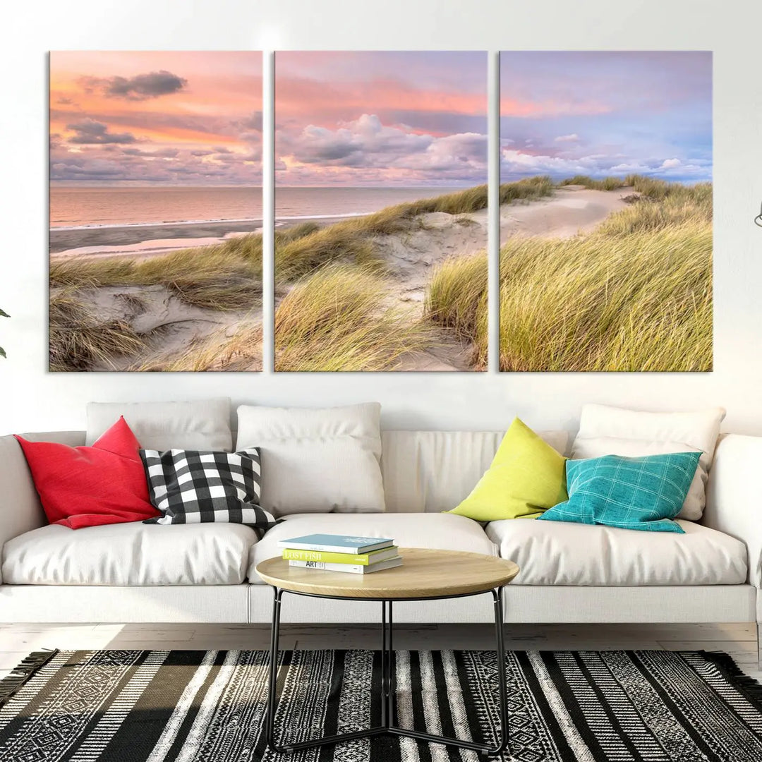 The Ocean Beach Wall Art Canvas Print Sunset Artwork captures a stunning sunset over sand dunes and grass, adding a touch of coastal charm to the room. This piece of wall art infuses any space with warmth and serenity.