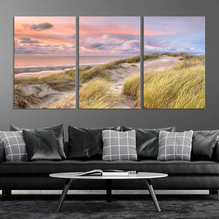 The Ocean Beach Wall Art Canvas Print Sunset Artwork captures a stunning sunset over sand dunes and grass, adding a touch of coastal charm to the room. This piece of wall art infuses any space with warmth and serenity.