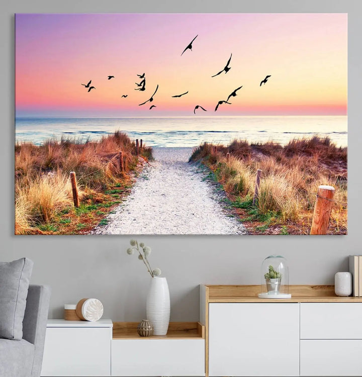 The "Ocean Pathway Sunset Canvas Art" beautifully depicts a tranquil coastal beach walk at sunset, featuring birds in flight across a vividly colorful sky. This piece is crafted using giclee print technology on canvas to ensure vibrant colors and exquisite detail for a relaxing wall art experience.