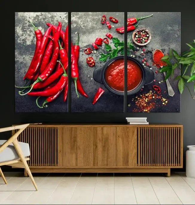 The Red Chili Peppers Wall Art Kitchen Artwork Canvas Print is a three-panel canvas showcasing red chili peppers, sliced chili, a pot of red sauce, and assorted spices on a dark background. It is of museum quality with a UV-protective coating to ensure its colors remain vibrant over time.