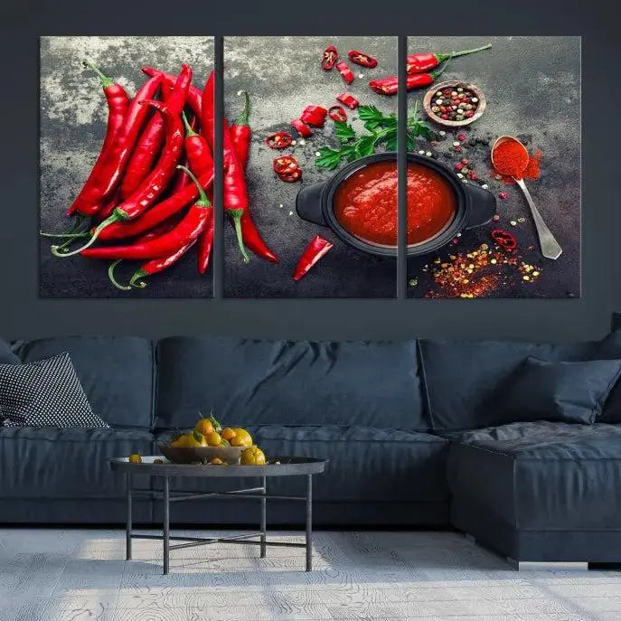 The Red Chili Peppers Wall Art Kitchen Artwork Canvas Print is a three-panel canvas showcasing red chili peppers, sliced chili, a pot of red sauce, and assorted spices on a dark background. It is of museum quality with a UV-protective coating to ensure its colors remain vibrant over time.