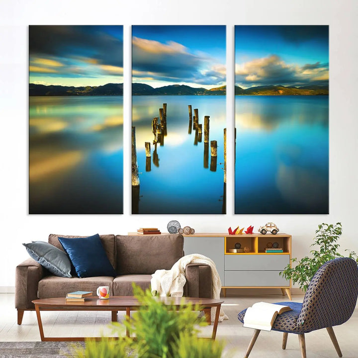 A triptych of a serene lake scene with old wooden pillars, blue sky, and clouds casts a coastal ambiance. The Sea Ocean Sunset Beach Wall Art Canvas Print reflects the soothing nature of the ocean.