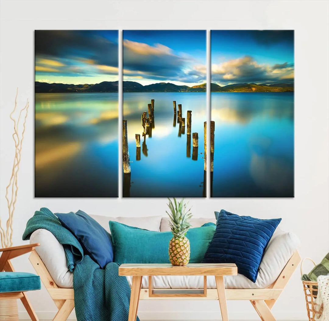 A triptych of a serene lake scene with old wooden pillars, blue sky, and clouds casts a coastal ambiance. The Sea Ocean Sunset Beach Wall Art Canvas Print reflects the soothing nature of the ocean.