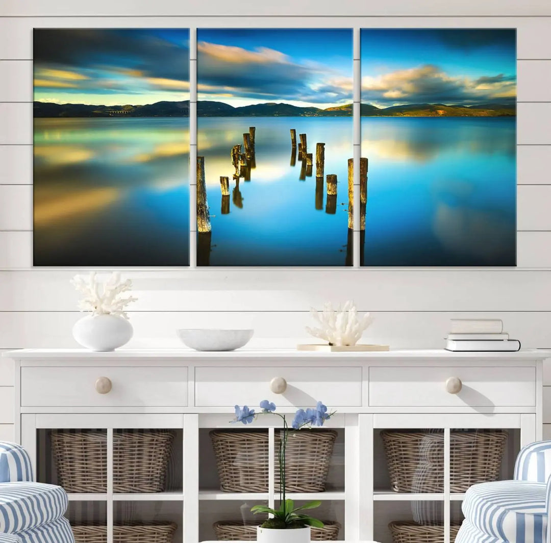 A triptych of a serene lake scene with old wooden pillars, blue sky, and clouds casts a coastal ambiance. The Sea Ocean Sunset Beach Wall Art Canvas Print reflects the soothing nature of the ocean.