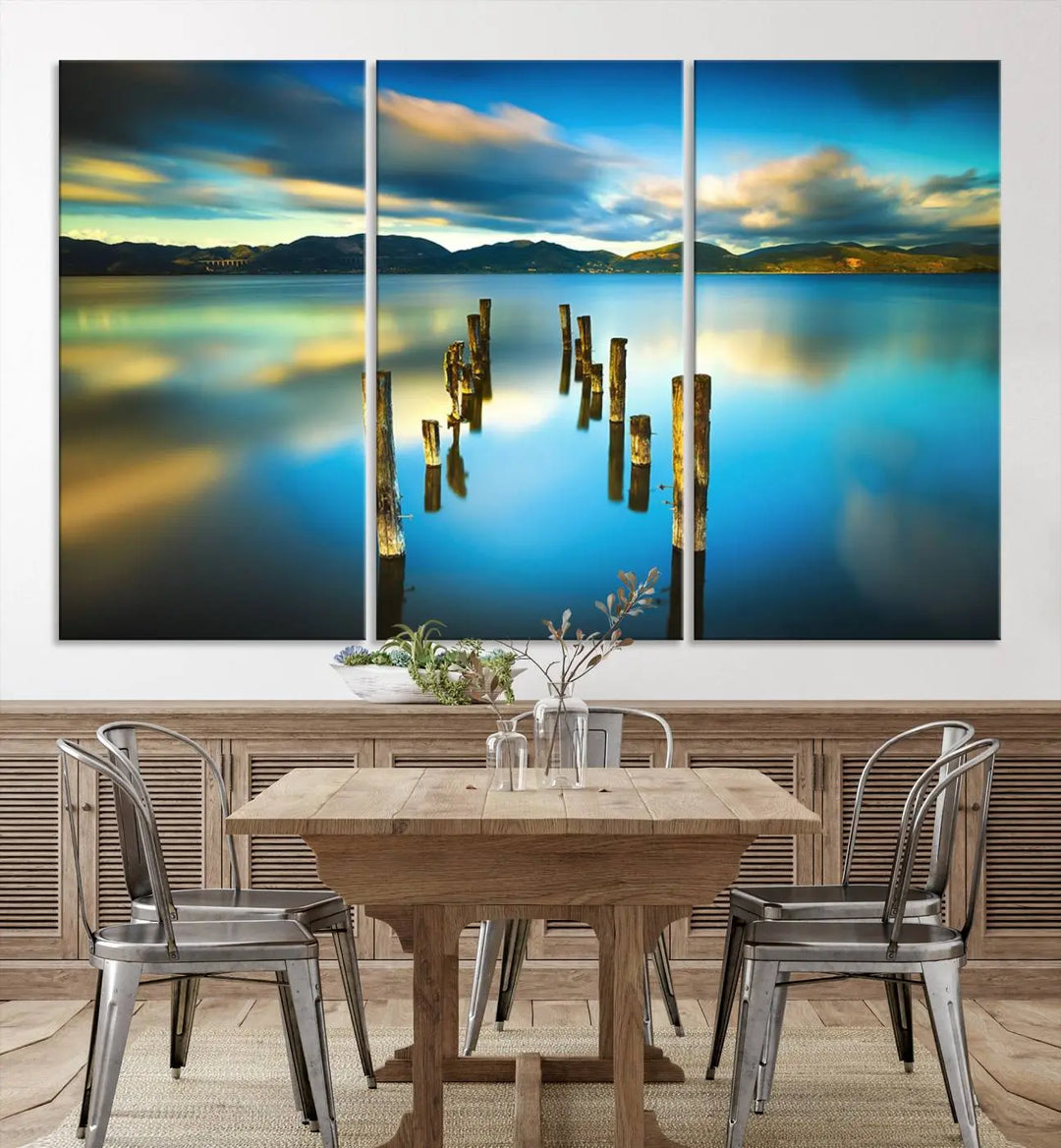 A triptych of a serene lake scene with old wooden pillars, blue sky, and clouds casts a coastal ambiance. The Sea Ocean Sunset Beach Wall Art Canvas Print reflects the soothing nature of the ocean.