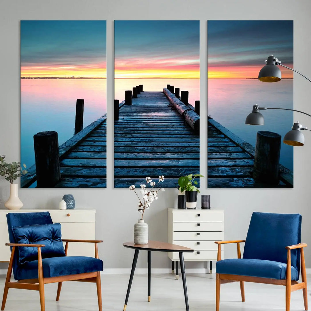 The "Sunset Pier Triptych Canvas Art," a giclee print known for its Canon print quality and gallery wrap finish, captures a wooden pier stretching into a tranquil lake at sunset beneath vibrant orange and blue skies. It makes an ideal centerpiece for coastal-themed art settings.