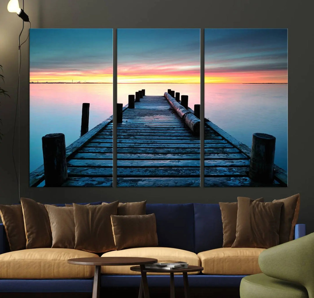 The "Sunset Pier Triptych Canvas Art," a giclee print known for its Canon print quality and gallery wrap finish, captures a wooden pier stretching into a tranquil lake at sunset beneath vibrant orange and blue skies. It makes an ideal centerpiece for coastal-themed art settings.