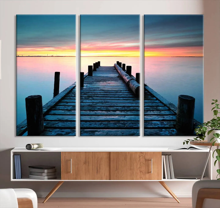 The "Sunset Pier Triptych Canvas Art," a giclee print known for its Canon print quality and gallery wrap finish, captures a wooden pier stretching into a tranquil lake at sunset beneath vibrant orange and blue skies. It makes an ideal centerpiece for coastal-themed art settings.