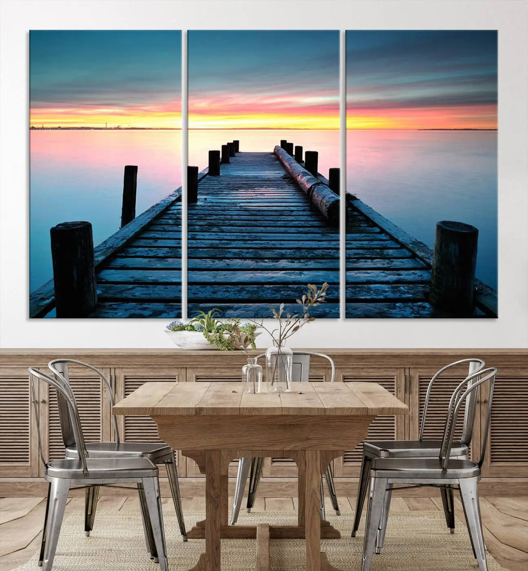 The "Sunset Pier Triptych Canvas Art," a giclee print known for its Canon print quality and gallery wrap finish, captures a wooden pier stretching into a tranquil lake at sunset beneath vibrant orange and blue skies. It makes an ideal centerpiece for coastal-themed art settings.