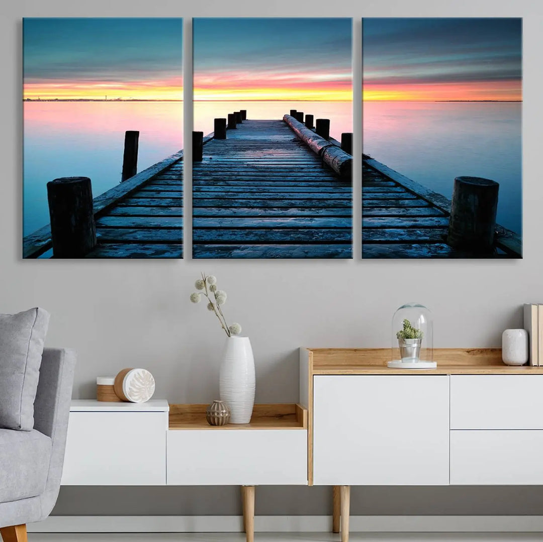 The "Sunset Pier Triptych Canvas Art," a giclee print known for its Canon print quality and gallery wrap finish, captures a wooden pier stretching into a tranquil lake at sunset beneath vibrant orange and blue skies. It makes an ideal centerpiece for coastal-themed art settings.