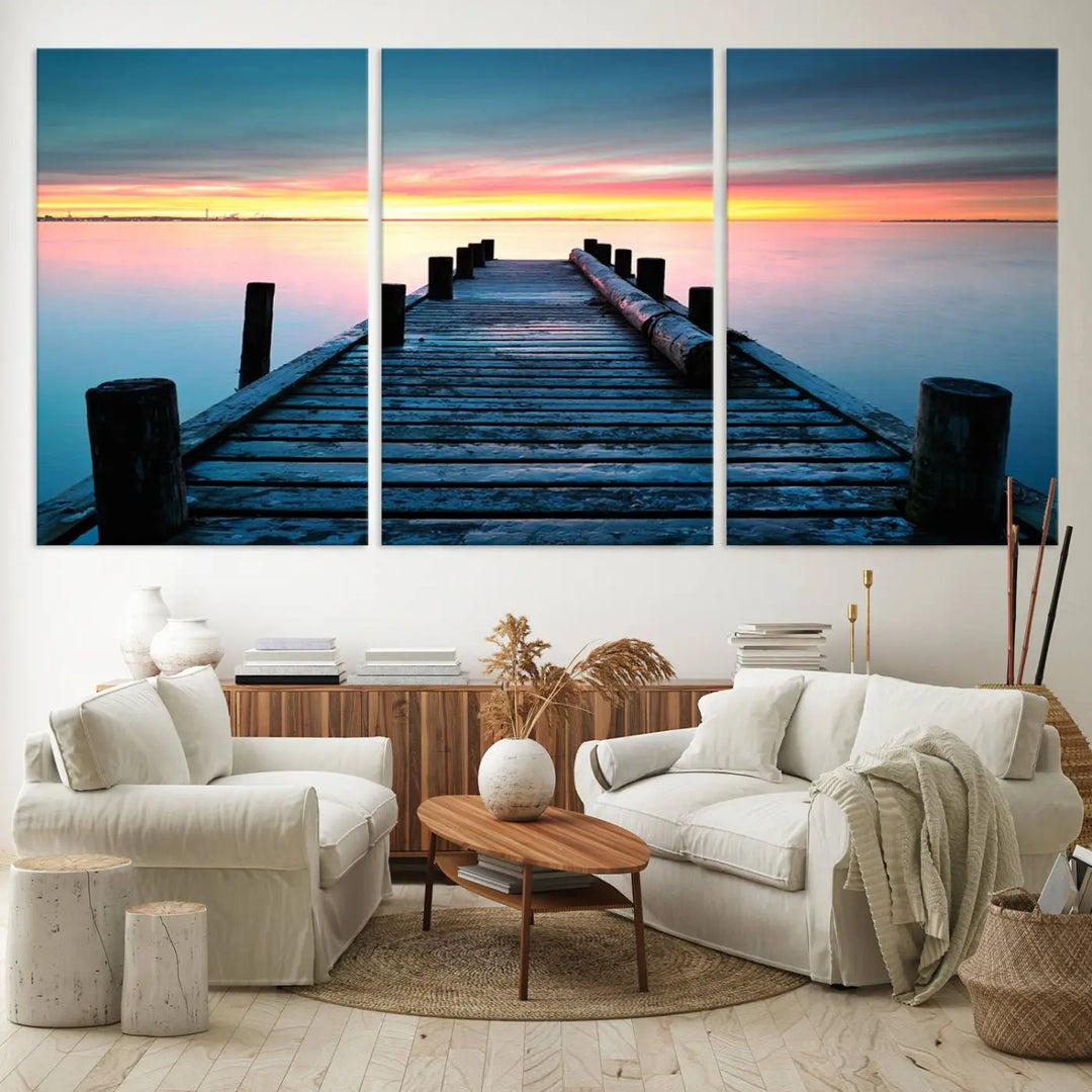 The "Sunset Pier Triptych Canvas Art," a giclee print known for its Canon print quality and gallery wrap finish, captures a wooden pier stretching into a tranquil lake at sunset beneath vibrant orange and blue skies. It makes an ideal centerpiece for coastal-themed art settings.