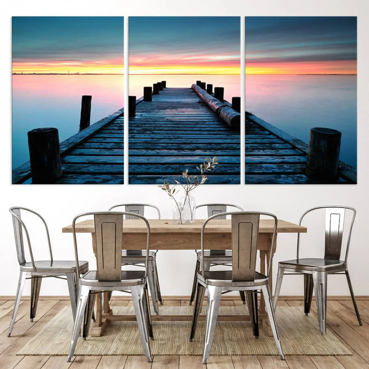 The "Sunset Pier Triptych Canvas Art," a giclee print known for its Canon print quality and gallery wrap finish, captures a wooden pier stretching into a tranquil lake at sunset beneath vibrant orange and blue skies. It makes an ideal centerpiece for coastal-themed art settings.