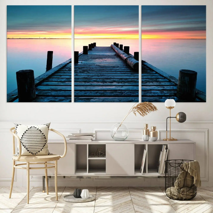 The "Sunset Pier Triptych Canvas Art," a giclee print known for its Canon print quality and gallery wrap finish, captures a wooden pier stretching into a tranquil lake at sunset beneath vibrant orange and blue skies. It makes an ideal centerpiece for coastal-themed art settings.