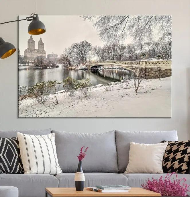 The Bow Bridge in Central Park Canvas Print features a snowy scene with a bridge and city buildings in the background. This museum-quality canvas comes with a UV-protective coating and is ready to hang.