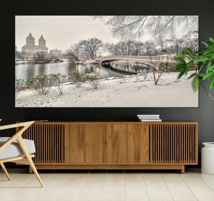 The Bow Bridge in Central Park Canvas Print features a snowy scene with a bridge and city buildings in the background. This museum-quality canvas comes with a UV-protective coating and is ready to hang.