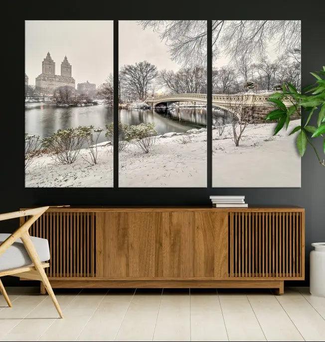 The Bow Bridge in Central Park Canvas Print features a snowy scene with a bridge and city buildings in the background. This museum-quality canvas comes with a UV-protective coating and is ready to hang.