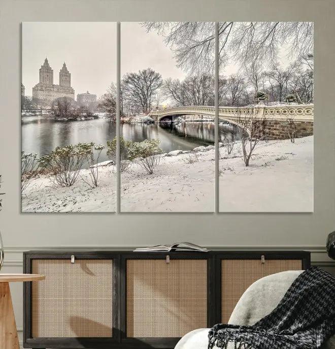 The Bow Bridge in Central Park Canvas Print features a snowy scene with a bridge and city buildings in the background. This museum-quality canvas comes with a UV-protective coating and is ready to hang.