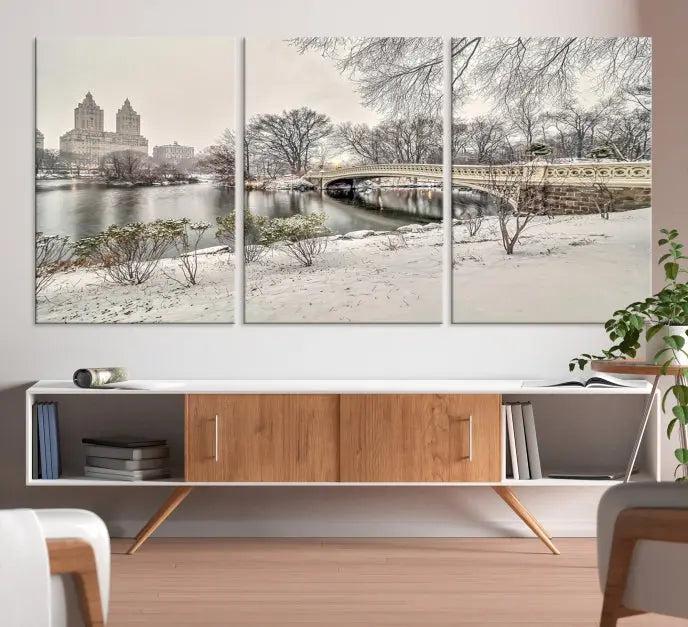 The Bow Bridge in Central Park Canvas Print features a snowy scene with a bridge and city buildings in the background. This museum-quality canvas comes with a UV-protective coating and is ready to hang.