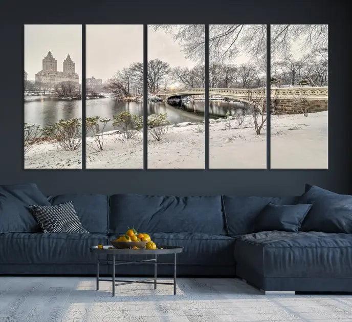 The Bow Bridge in Central Park Canvas Print features a snowy scene with a bridge and city buildings in the background. This museum-quality canvas comes with a UV-protective coating and is ready to hang.