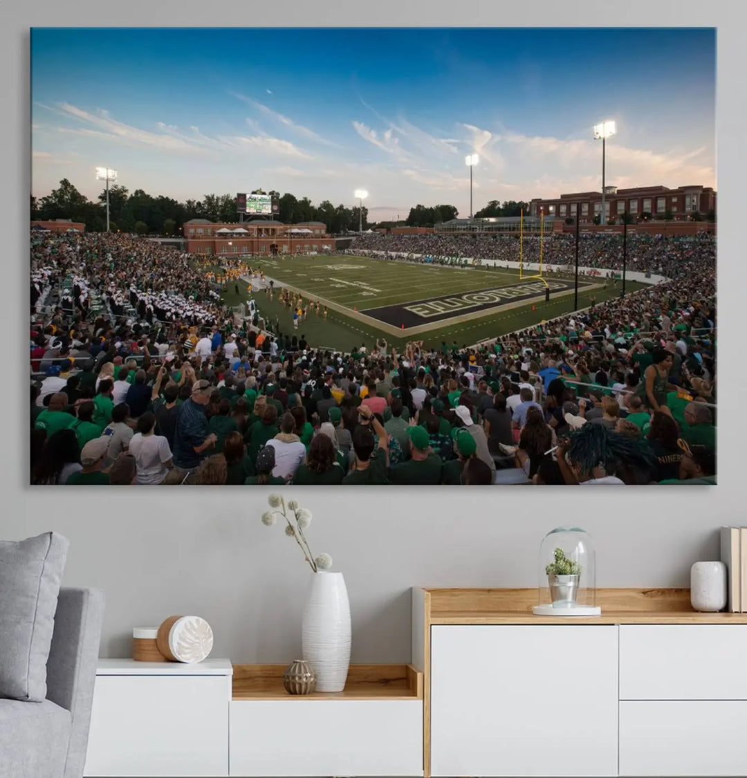 Above the dining table hangs a premium canvas triptych depicting Charlotte Jerry Richardson Stadium, home of the University of Charlotte 49ers Football Team. The scene is alive with a nighttime atmosphere, bustling with a large crowd under bright stadium lights. This stunning art piece is expertly handmade in the USA and adds vibrant energy to your living space.