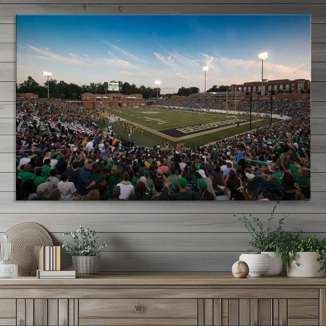 Above the dining table hangs a premium canvas triptych depicting Charlotte Jerry Richardson Stadium, home of the University of Charlotte 49ers Football Team. The scene is alive with a nighttime atmosphere, bustling with a large crowd under bright stadium lights. This stunning art piece is expertly handmade in the USA and adds vibrant energy to your living space.